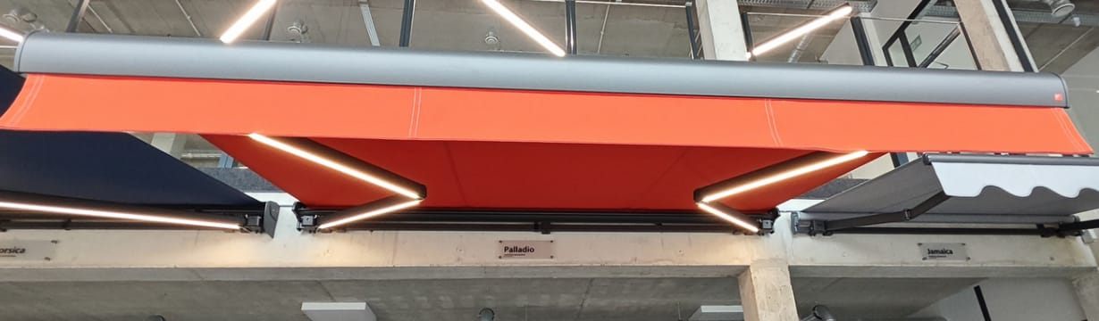 Markīze ar LED apgaismojumu svirās: vakarā terase ir izgaismota bez atsevišķiem gaismekļiem. Izvelkama markīze ar sarkanu audumu un LED apgaismojumu sviras, skats no apakšas — FENSTORA, Rīga