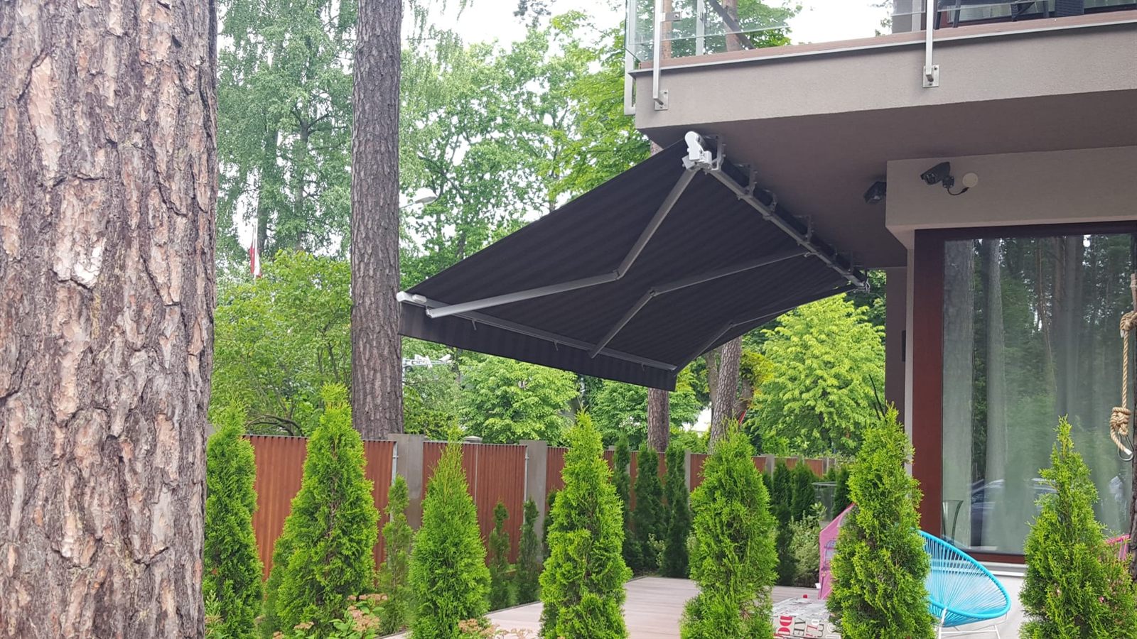 Pie baseina: antracīta markīze notur ēnu pār atpūtas zonu. Antracīta izvelkama markīze pie baseina starp priedēm zemesgabalā — FENSTORA, Rīga