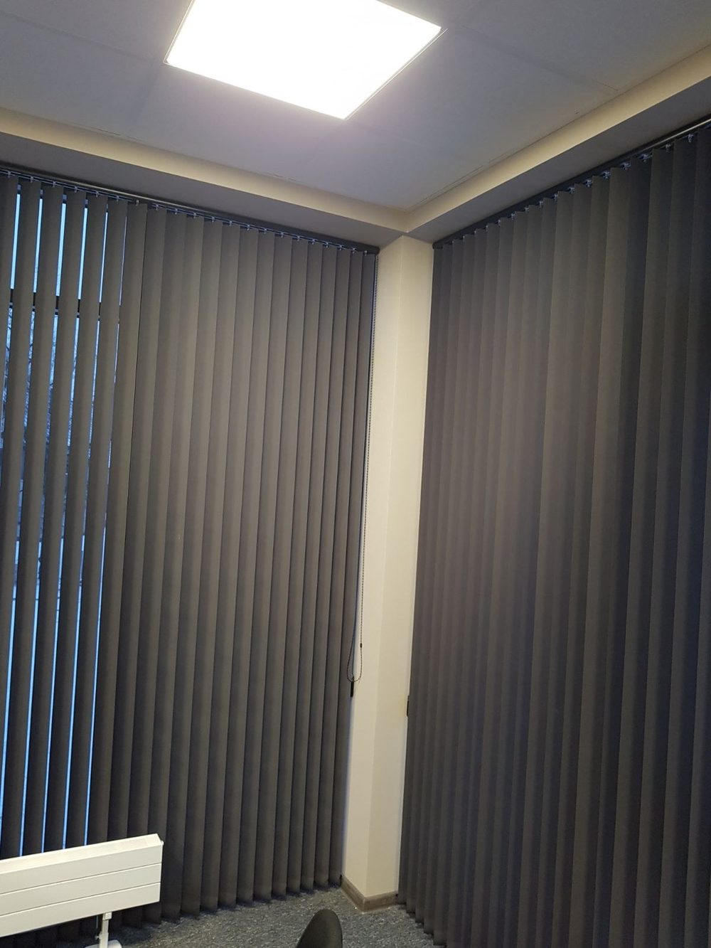Pelēkas vertikālās žalūzijas uz biroja stūra logiem