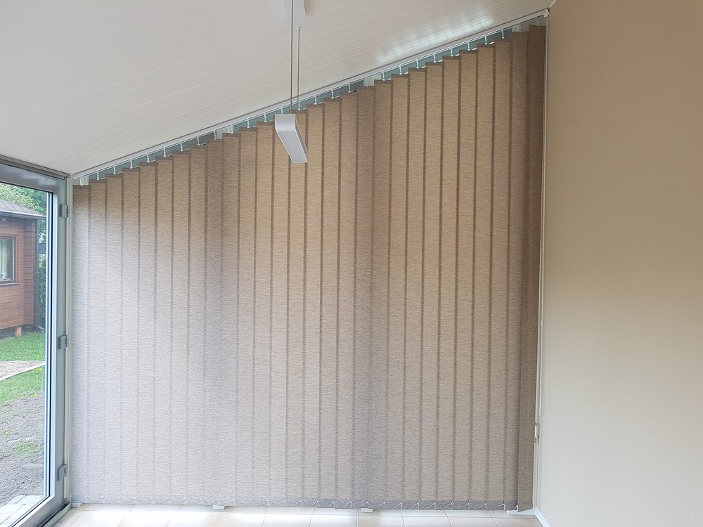 Bēšas vertikālās trapeces žalūzijas uz verandas slīpā loga